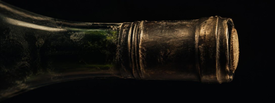 Extreme close-up of a sealed bottle neck lying on its side: the capsule seam and crimp lines under raking light, the edge of the cork just visible.
