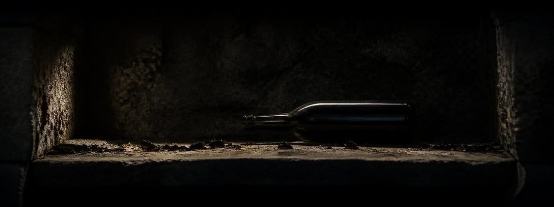 An empty stone vault niche: one unlabelled bottle lying on its side among scattered dark glass, under a single grazing light.