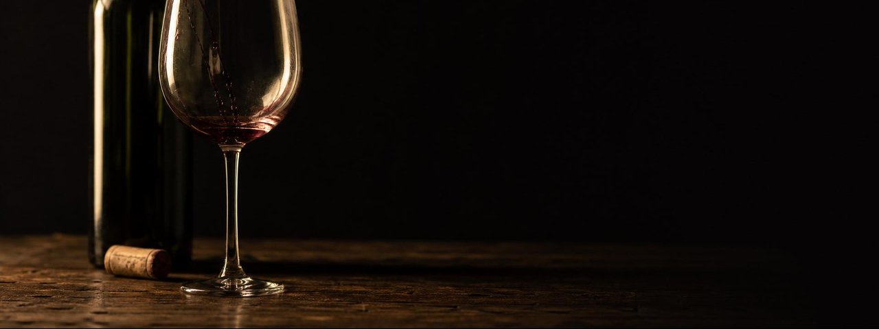 Una copa vac&iacute;a con las &uacute;ltimas huellas de vino tinto, una botella sin etiqueta y un corcho sobre una mesa oscura.