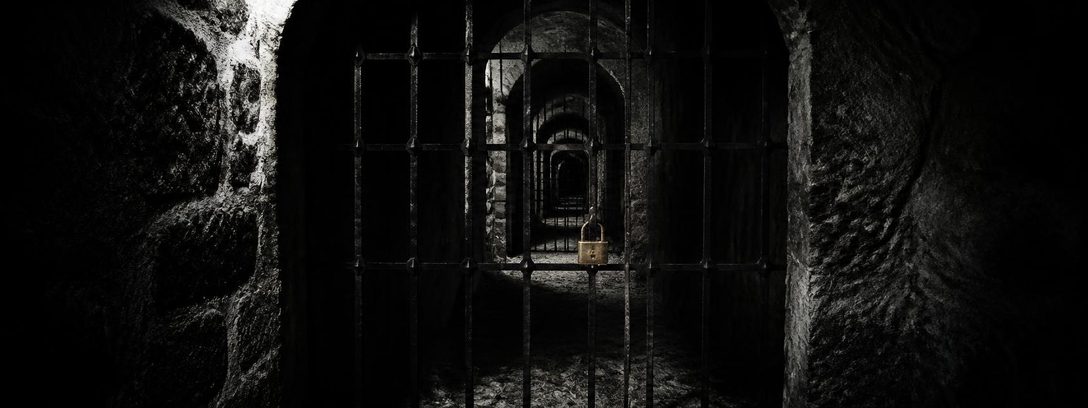 A dark stone cellar corridor with a sequence of iron gates receding into darkness; the nearest is closed with a brass padlock caught by the only light.