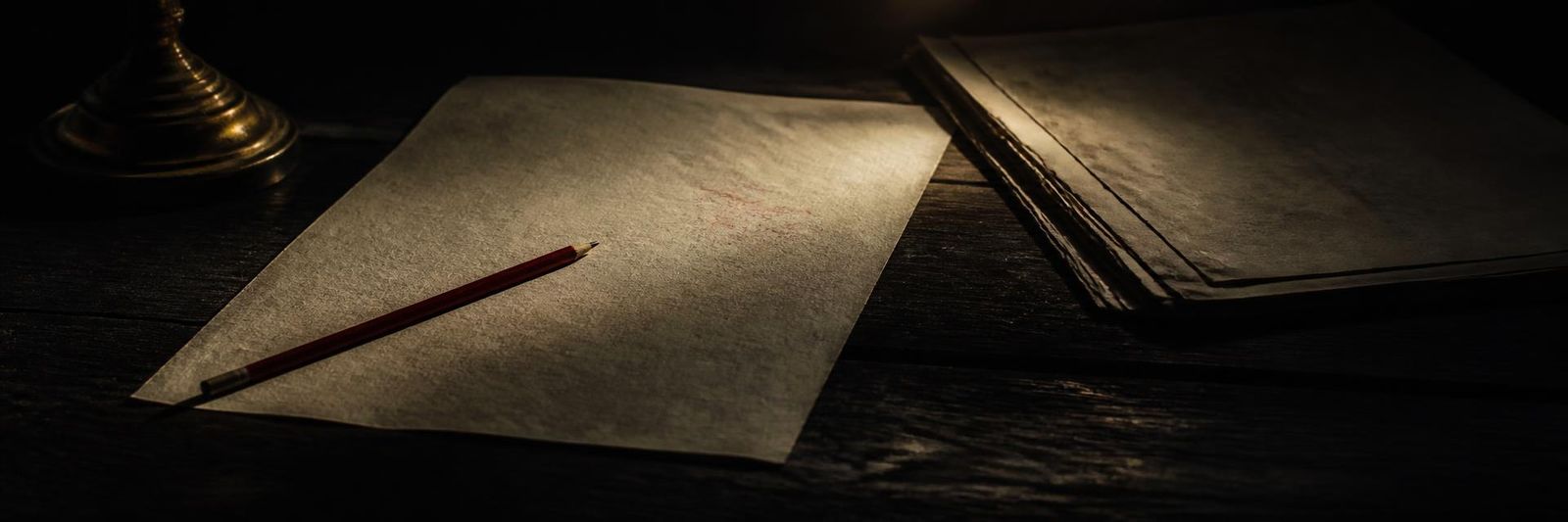 A sheet of paper on dark wood with a red pencil across it and a stack of older papers beside